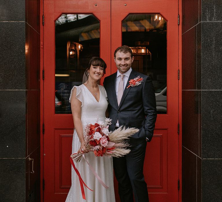 The bride in a sparkling glittery wedding dress with the groom in a navy suit in front of venue to intimate wedding 