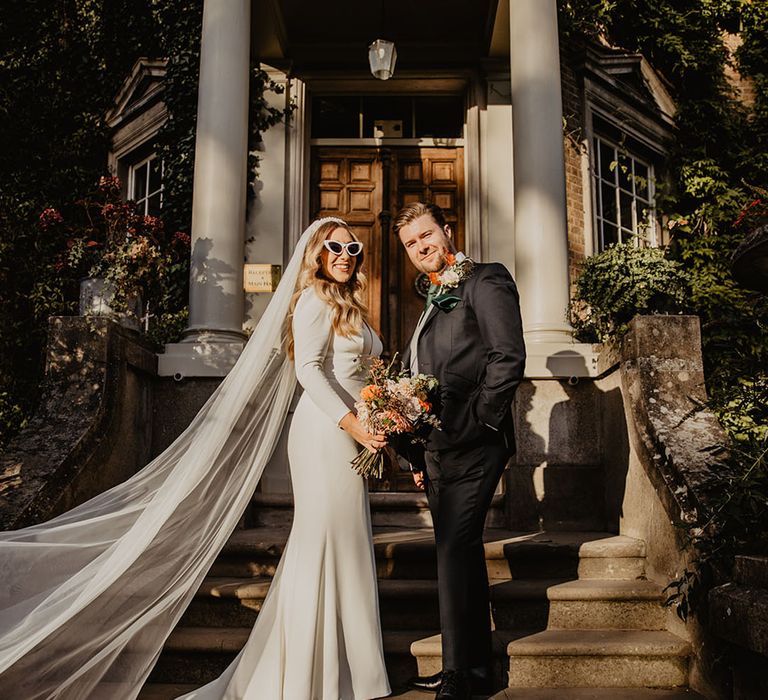 Bride in fitted wedding dress with retro sunglasses and pearl headband standing with the groom 