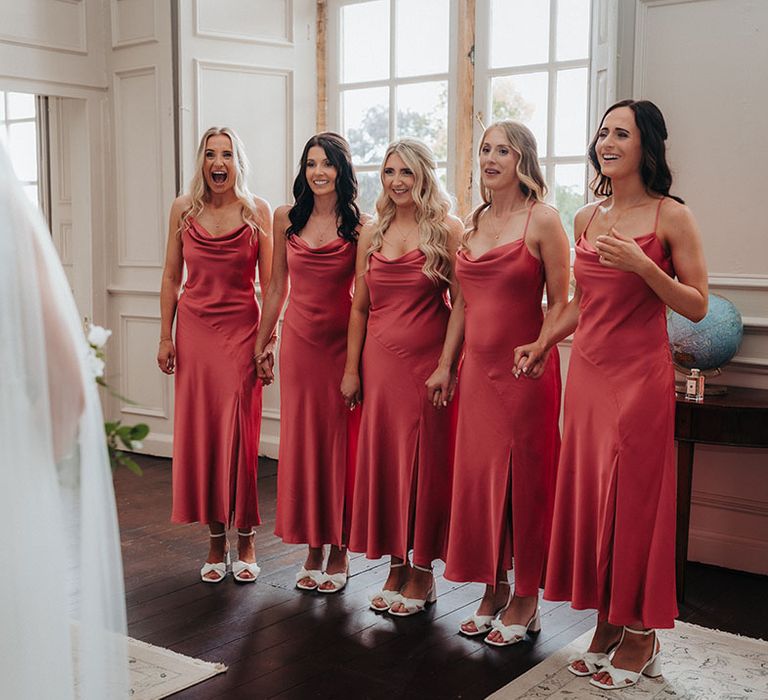 Bridesmaids in coral satin cowl bridesmaid dresses get their first look at the bride in her wedding dress 