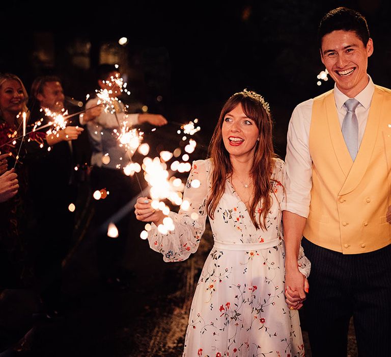 Groom in yellow waistcoat and blue tie walks with the bride waving sparklers for their sparkler send off 