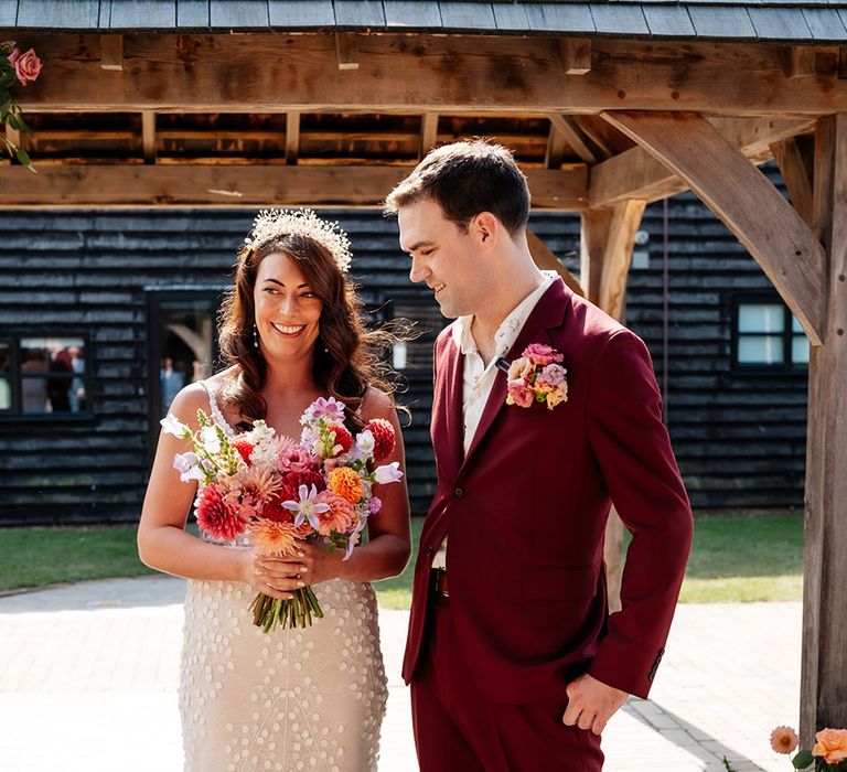 Groom in burgundy wedding suit with the bride in a Made With Love wedding dress carrying a colourful wedding bouquet 