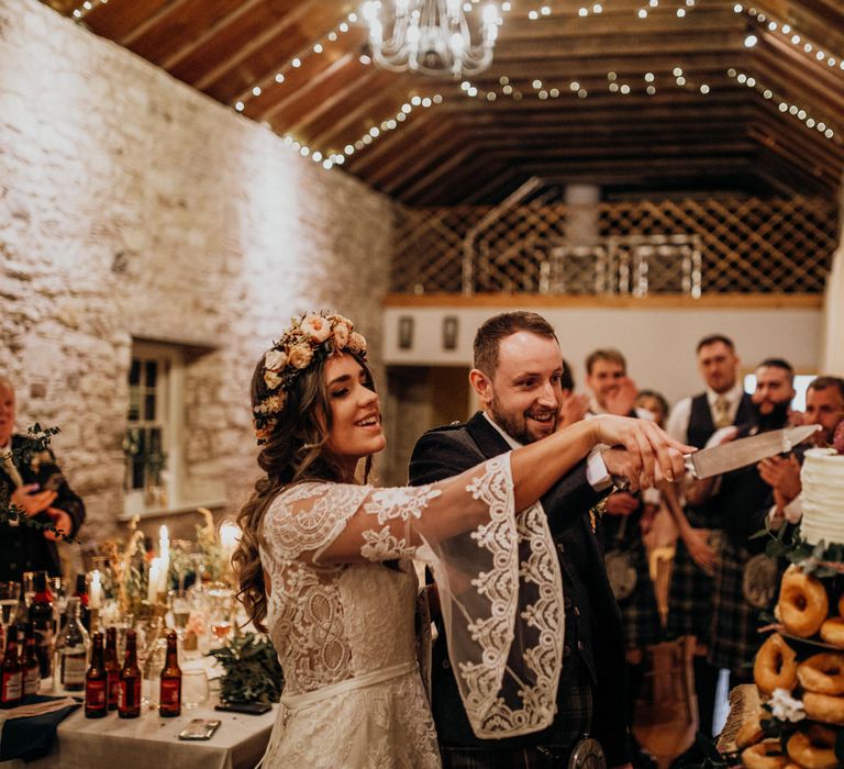 Boho bride in rose flower crown and groom in tartan cut doughnut cake at highland wedding in Glencoe