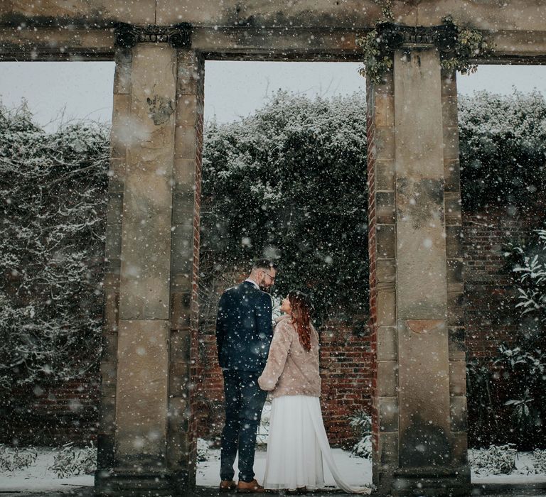 Bride in fur coat holding dried flower bouquet holds hands with groom in navy suit in the snowy grounds at Cannon Hall