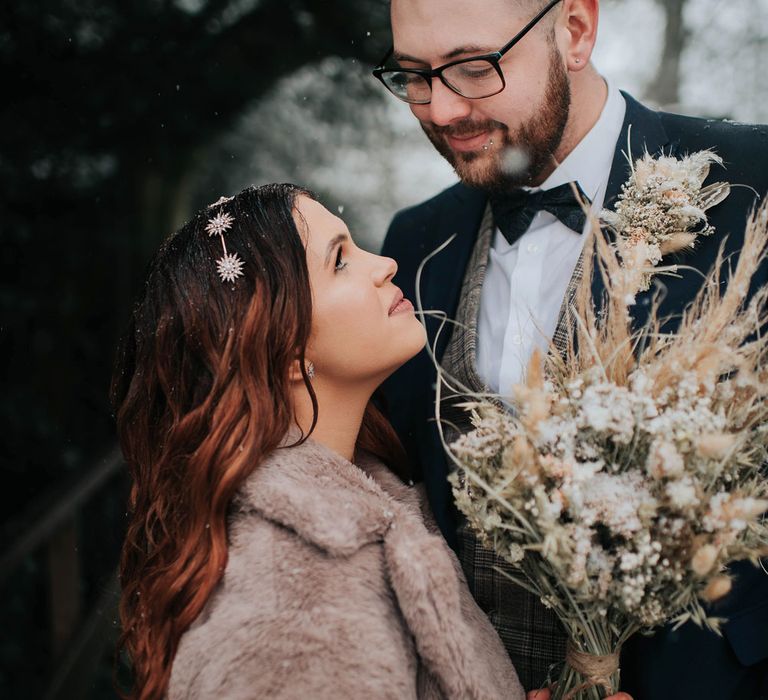 Bride in fur coat and celestial headband holding dried flower bouquet smiles up at groom in bow tie and navy suit