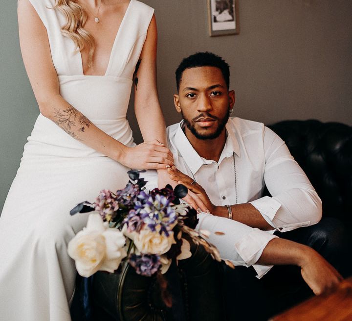 Portrait of a stylish bride and groom with the groom in leather trousers and white shirt sitting on a sofa and the bride on the arm in a crown and fitted dress