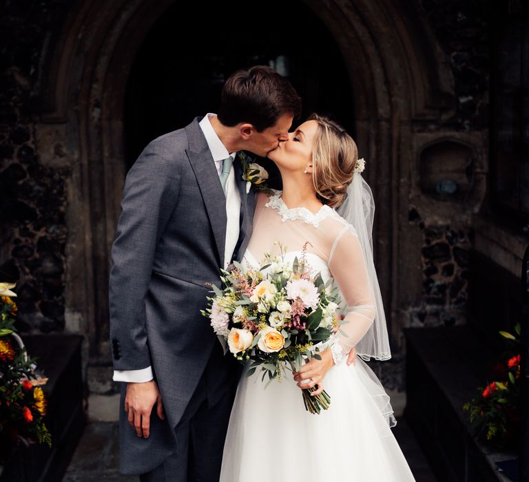 Bride & groom kiss outside church whilst bride holds classic bouquet