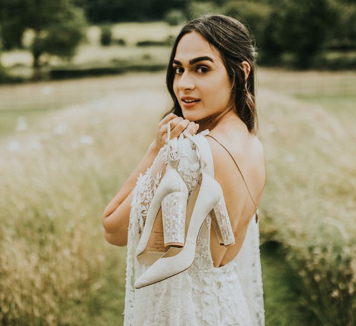 Bride in a lace wedding dress slinging her mother of pearl block heel shoes over her shoulder 