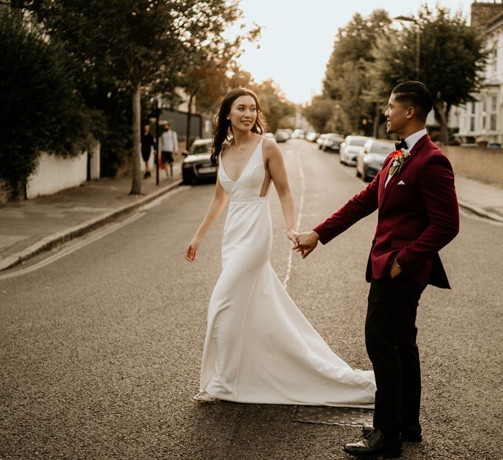 Bride in a fitted wedding dress with puddle train holding hands with her groom in a dark red blazer crossing the road in Clapton 