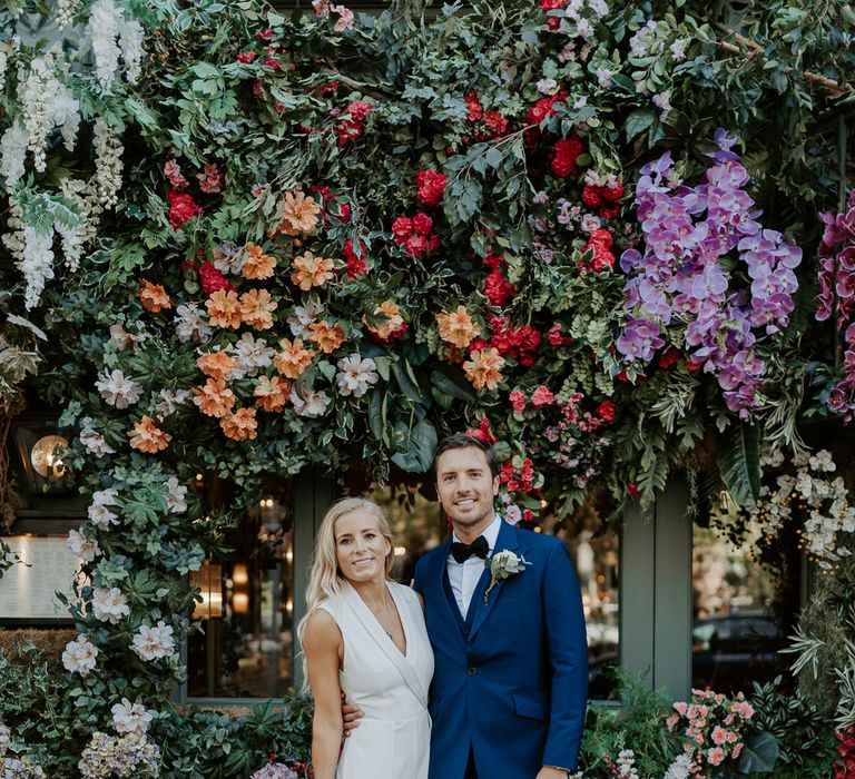 Bride & groom stand in front of colourful flower wall after wedding ceremony