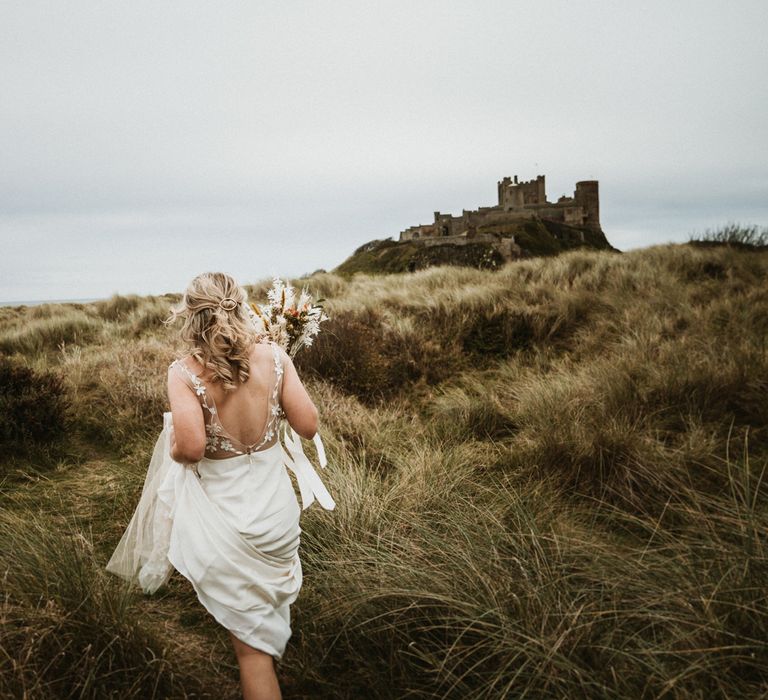 Bride walks through the grass whilst holding up dress with Bamburgh Castle in the background