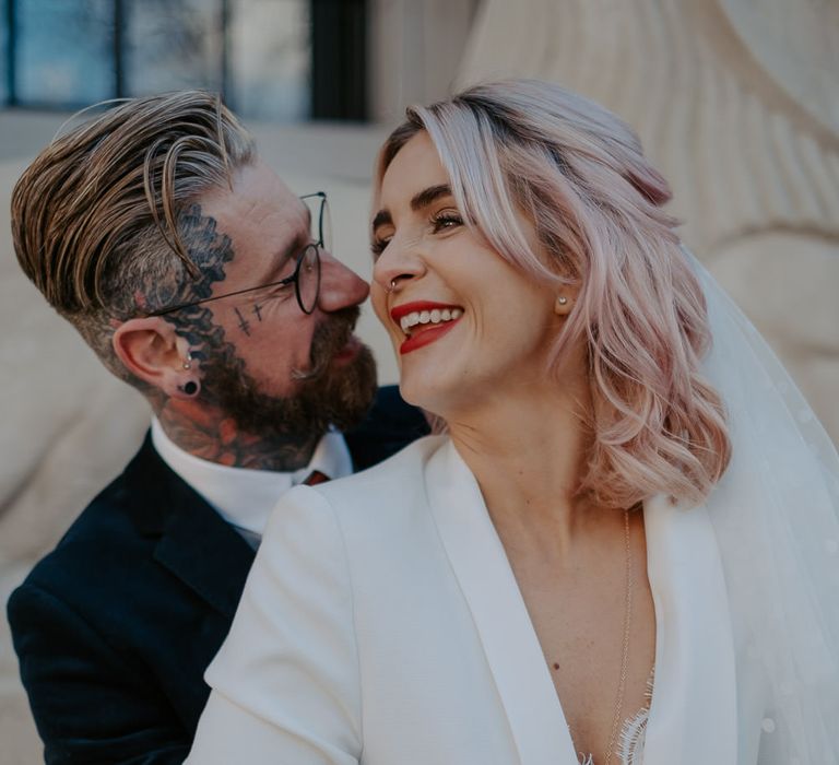 Groom with tattoos wraps arms around bride in white suit with pink hair and veil