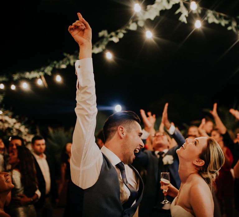 Bride and groom dancing in front of guests, the groom's arm in the air and the bride holding champagne