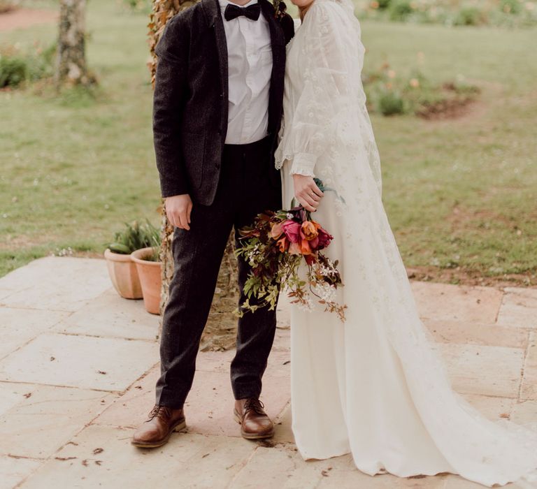 Bride in white Charlie Brear wedding dress with slit and chapel length daisy applique veil holding multicoloured wedding bouquet stands arm-in-arm with groom in grey tweed suit at garden party wedding in Devon