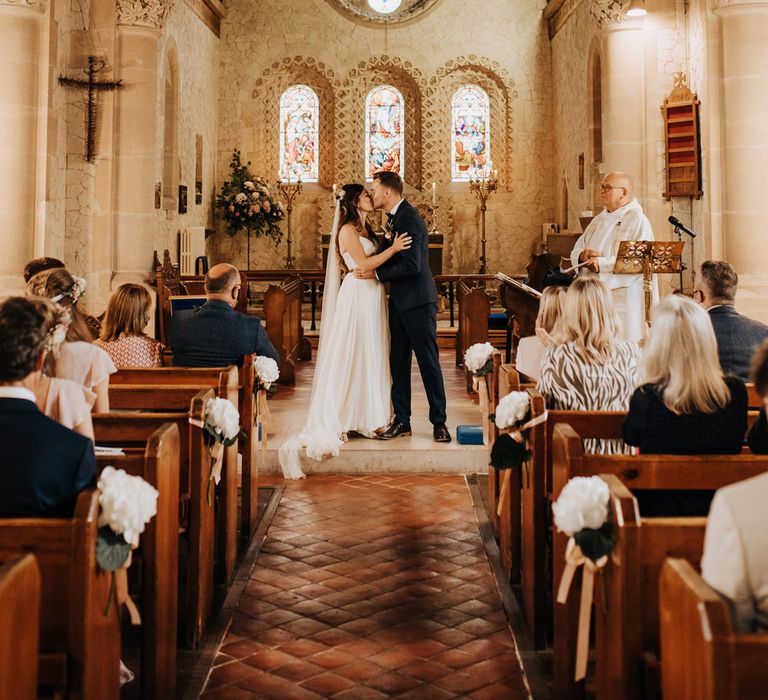 Bride in white cami dress with train and veil kisses groom in blue suit at the altar during wedding ceremony