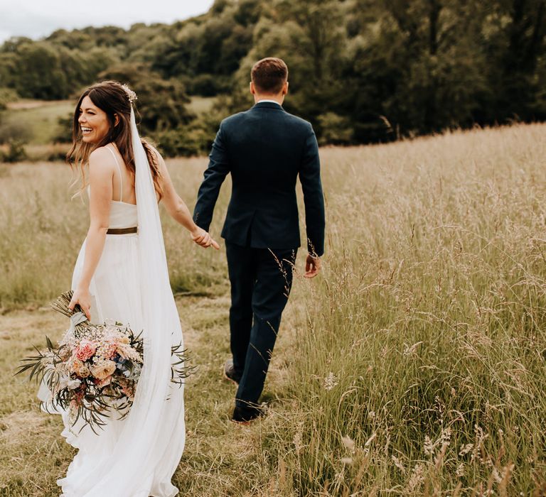 Bride in white dress and long veil holding multi coloured wedding bouquet walks into field hand-in-hand with groom in blue suit at garden wedding reception