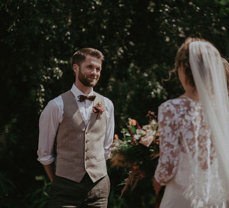 Groom in light brown waistcoat with bowtie, buttonhole and white shirt with rolled up sleeves smiles at bride in white Rime Arodaky wedding dress with mesh floral top during wedding ceremony with burnt orange wedding theme