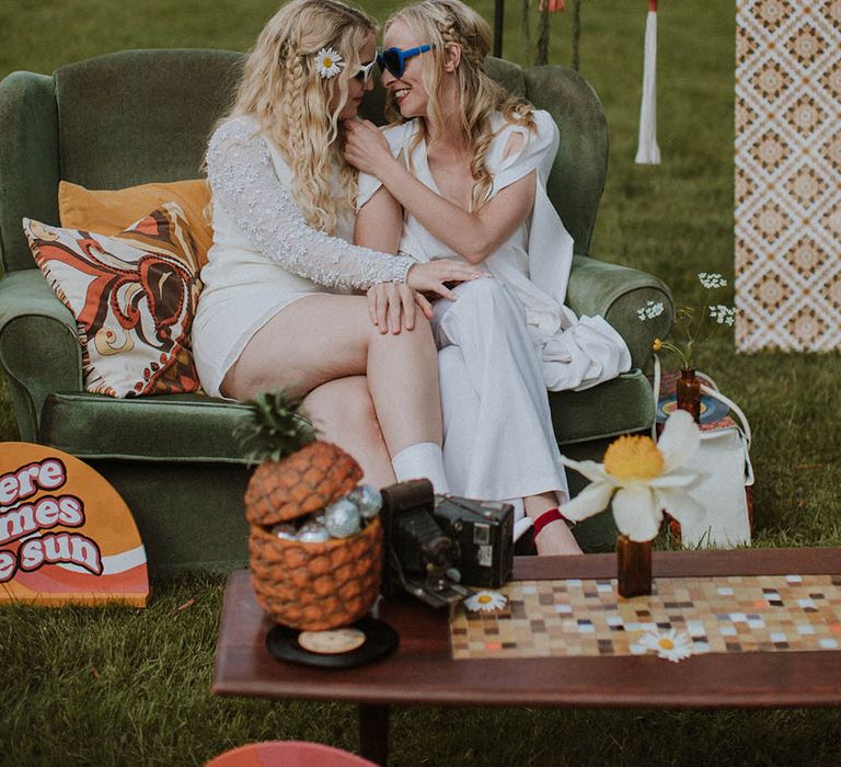 Two brides in sunglasses embracing on a green velvet loveseat at sixties wedding 