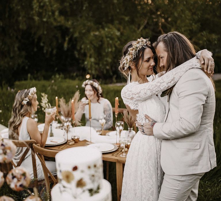 Groom in a beige suit embracing his boho bride in a lace wedding dress and dried flower crown by their outdoor table scape 