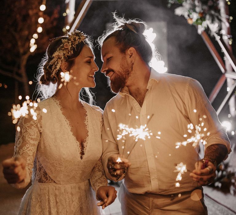 Boho bride in a lace wedding dress with dried flower crown and groom in a white shirt and beige trousers holding sparklers under a naked tipi 