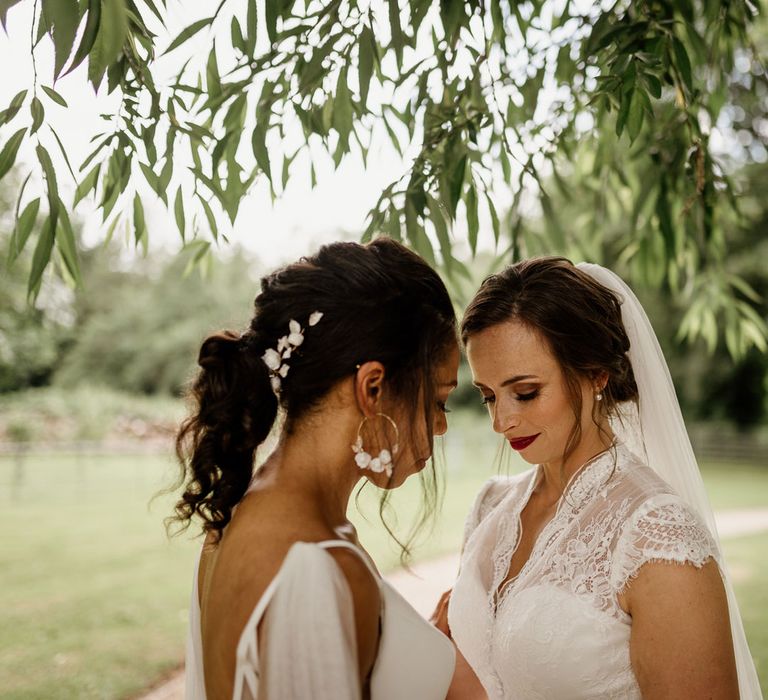 Bride with curled ponytail and hoop earrings stands with bride in lace capped sleeve wedding dress and veil in the grounds of The West Mill Derby after wedding ceremony