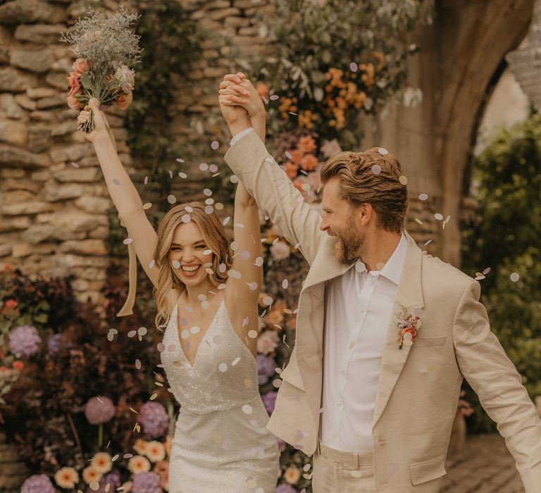 Groom in a beige suit and white shirt holding hands with his bride in a sparkly wedding dress as they wavy hands and celebrate as confetti is thrown over them