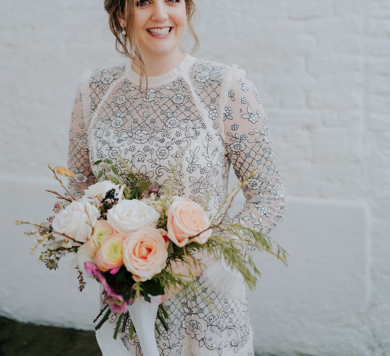 Bride smiles brightly as she looks toward the camera and holds pastel floral bouquet whilst wearing Needle And Thread wedding dress