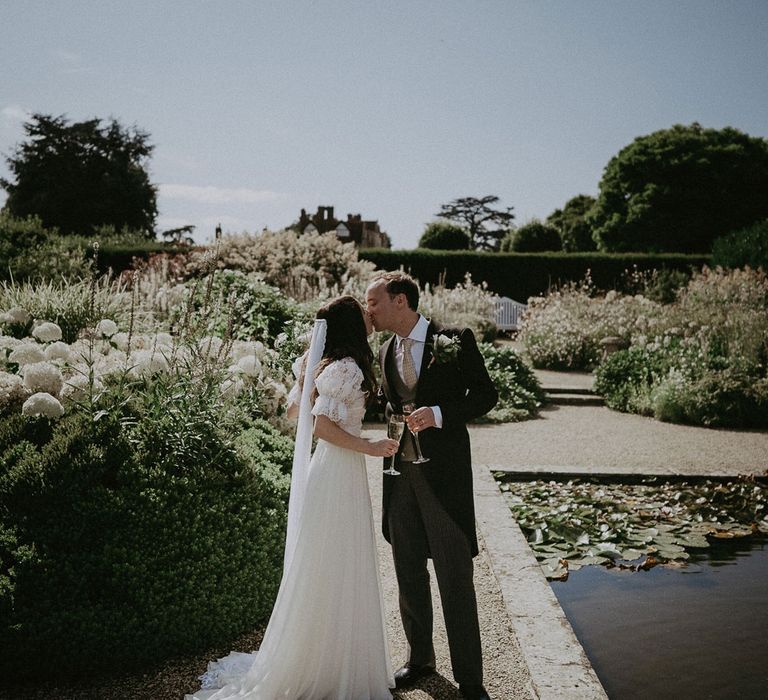 Bride in white lace puffed sleeve Daarlana wedding dress and bespoke veil stands in Loseley Park grounds kissing groom in morning coat and green waistcoat both holding sparkling wine glasses at summer wedding reception in Surrey