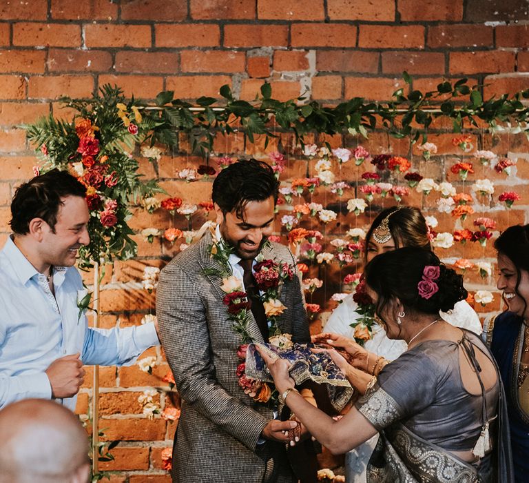 A groom in a grey check suit smiles during a ritual at his wedding ceremony.