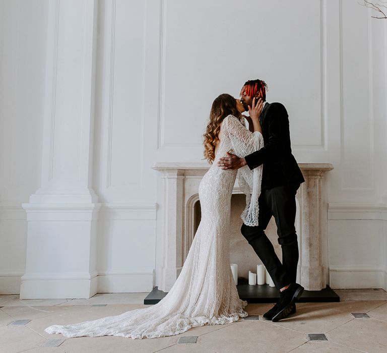 Stylish groom in a black velvet jacket and suede shoes kissing his bride in an embellished long sleeve wedding dress by the fireplace at Bylaugh Hall 