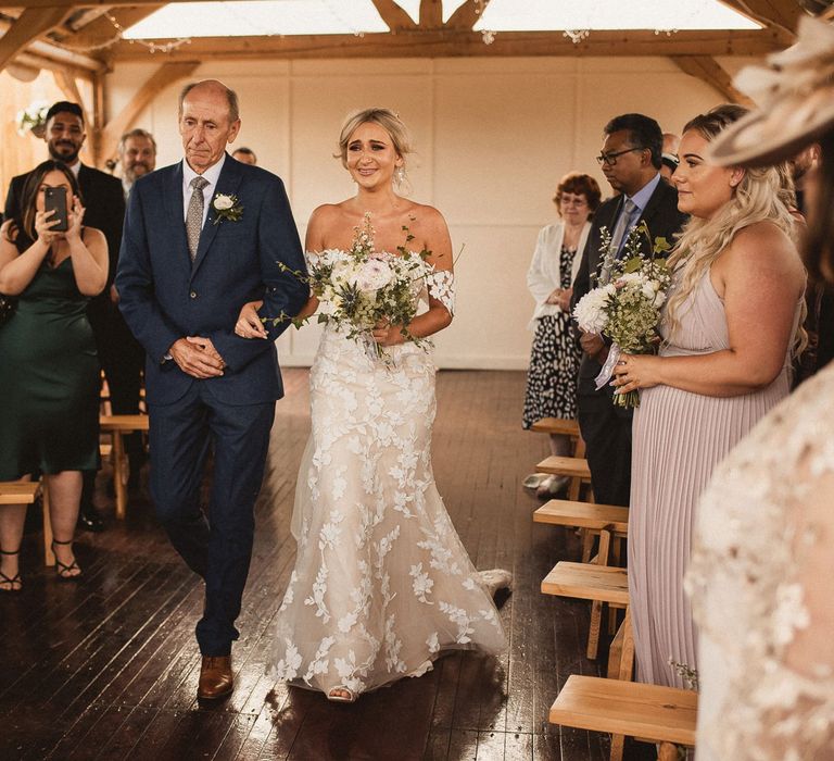 Smiling bride in off the shoulder Enzoani wedding dress holding white and green floral bouquet walks down the aisle holding arm of father of the bride in blue suit with white floral buttonhole at Inkersall Grange Farm wedding