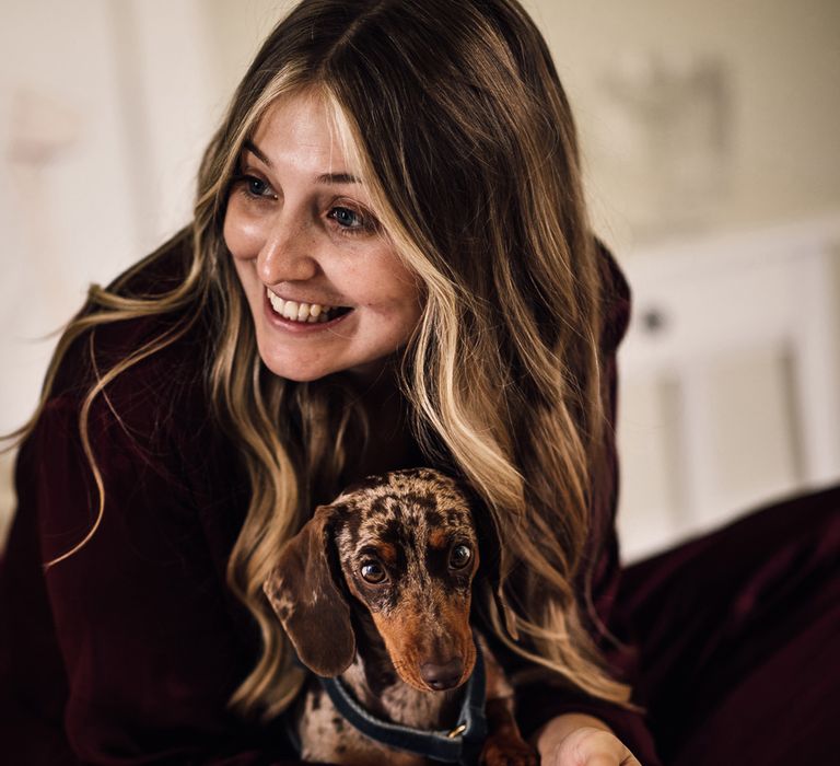 Bride in burgundy pyjamas with curled hair hugs miniature daschund before church wedding ceremony in Cornwall