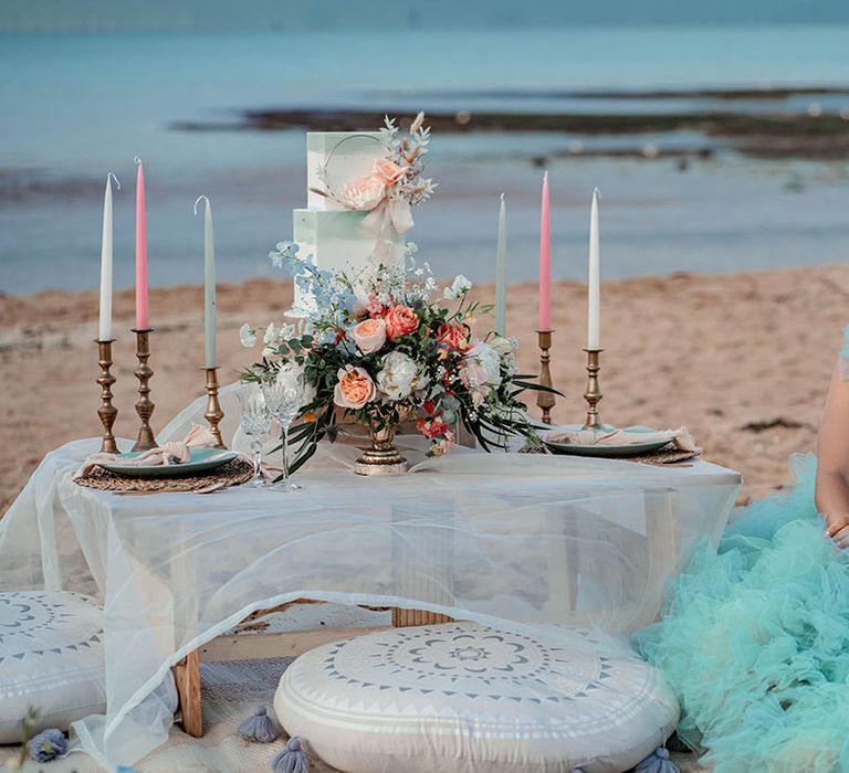 Bride in a mint green tulle wedding dress sitting at her relaxed beach wedding reception decorated with pastel flowers, decor and cakes 