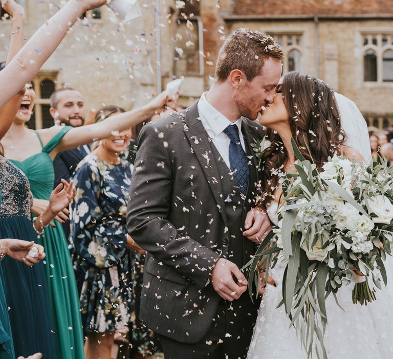 Bride in Pronovias wedding dress and veil holding white and green bridal bouquet kisses groom in grey three piece suit, blue tie and rose buttonhole as guests throw confetti at Notley Abbey wedding