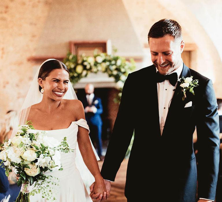 Bride & groom walk hand in hand down the aisle as bride wears Halfpenny London veil