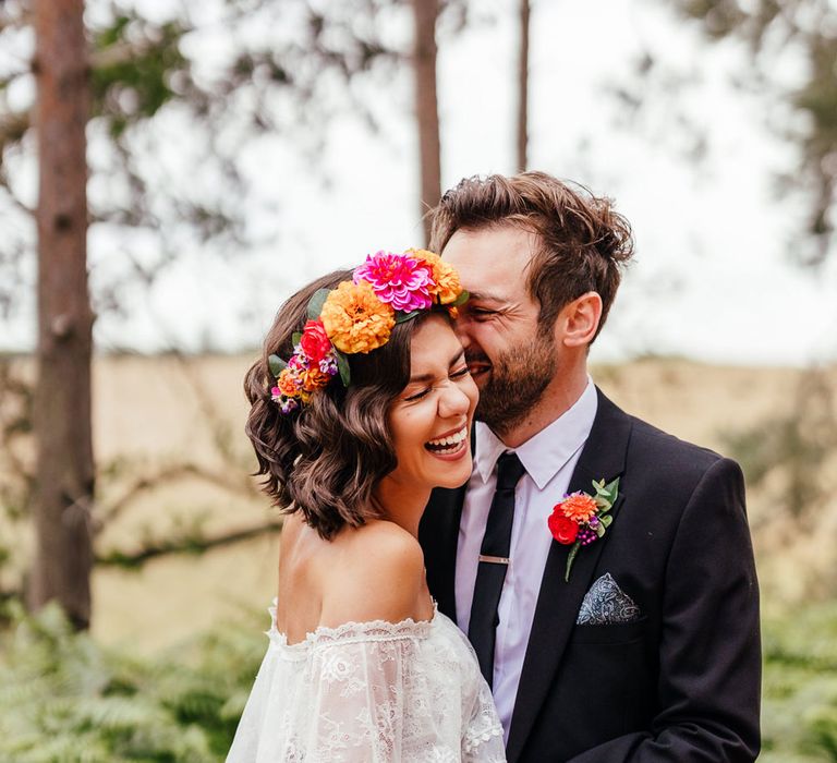 Bride in off the shoulder Grace Loves Lace wedding dress and flower crown holding multicoloured wedding bouquet laughs as she hugs groom in black suit and colourful floral buttonhole in woodland