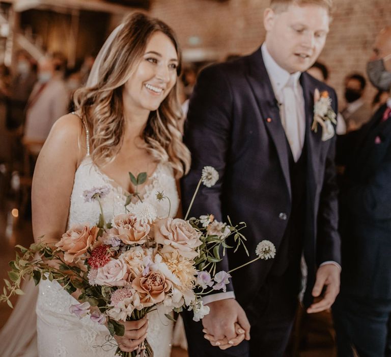 Bride holds floral bouquet in pastel colours as she walks with her groom