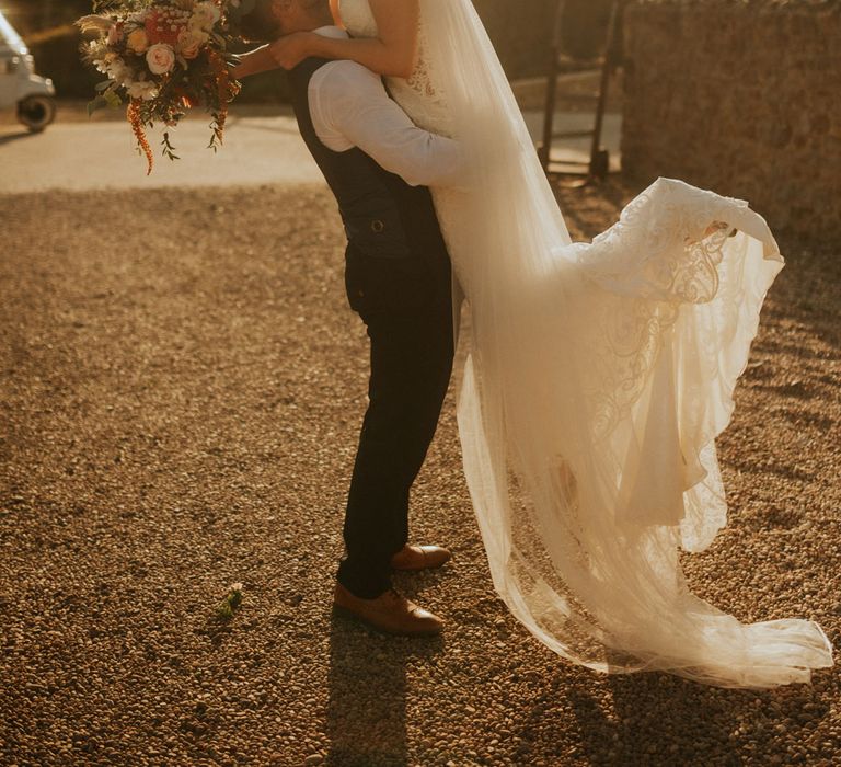 Groom in white shirt and waistcoat picks up bride in lace wedding dress with train and veil holding mixed bridal bouquet during golden hour