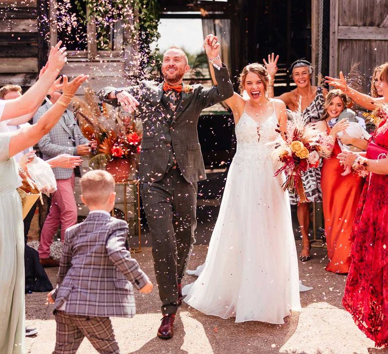 Bride in lace wedding dress with train holding mixed dried floral boho bouquet holds up arm with groom in green three piece suit and pattered shirt as guests throw confetti