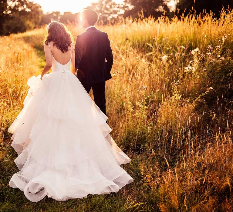 Bride in low back wedding dress with large tulle skirt and button detailing walks through field with groom in dark suit during golden hour