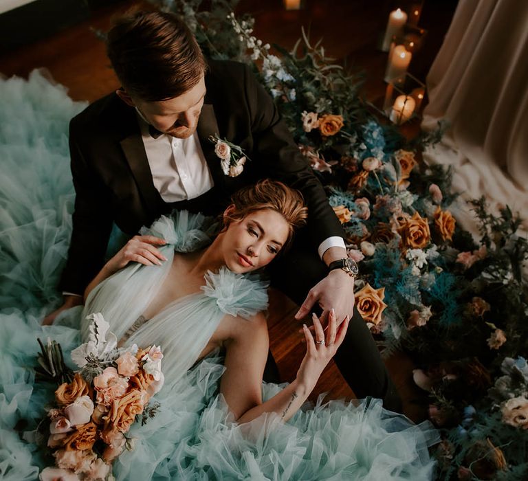 Bride in a light blue ruffle wedding dress laying in her grooms lap surrounded by cafe au late roses, pink orchids, ranunculus and carnations floral arrangement 