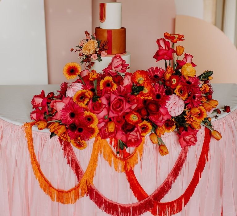 Wedding cake table with pink tablecloth and geometric backdrop and vibrant red floral arrangement and tassels 