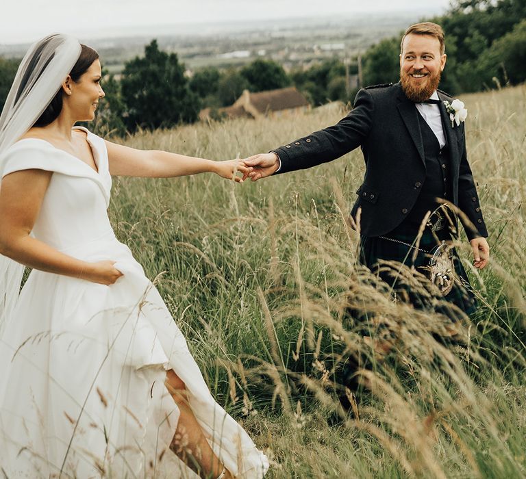 Groom in a tartan kilt holding hands with his bride in a Suzanne Neville princess wedding dress in field 