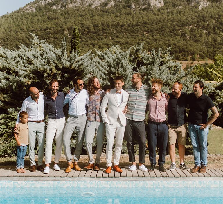 groom in beige suit and pink bow tie smiles with groomsmen in front of a pool in Mallorca