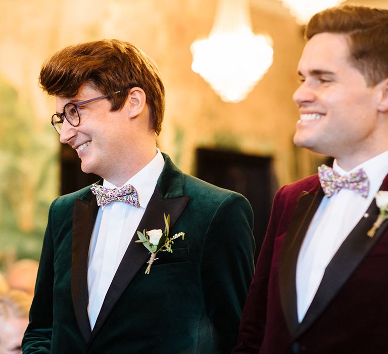 Two grooms in Burgundy and forest green velvet suit jackets with floral bow ties and white rose boutonnière