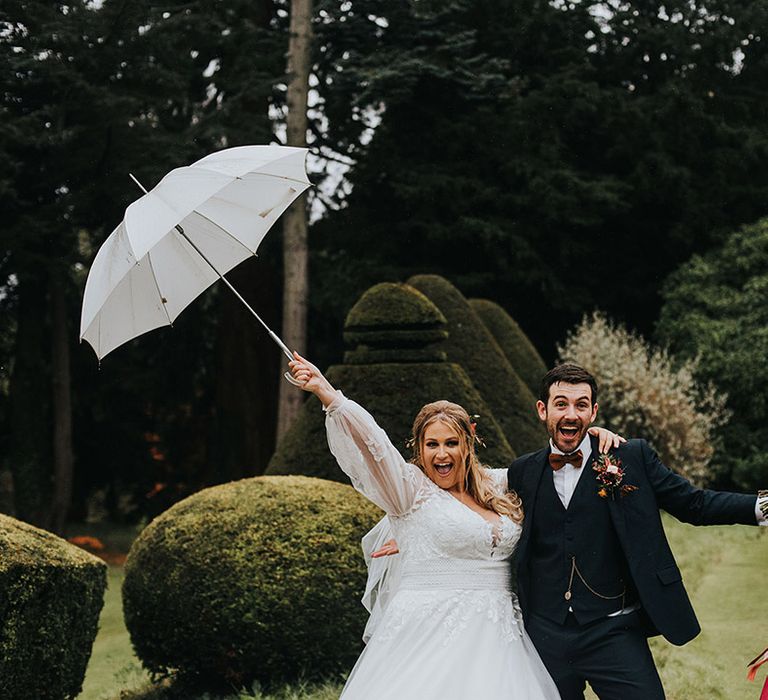 Rainy autumn wedding with bride in a princess wedding dress with long sleeves waving a white umbrella in the air and the groom in a navy suit holding a Burgundy bouquet 