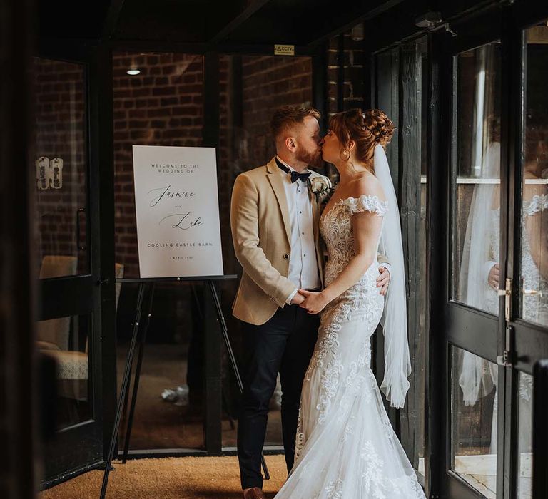 Bride and groom kiss next to black and white welcome wedding sign with personalised details