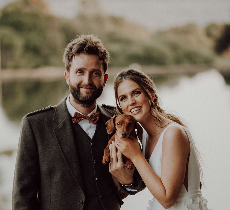 Groom in orange paisley bow tie with bride in Savin London wedding dress holding their dachshund pet, Ruby between them