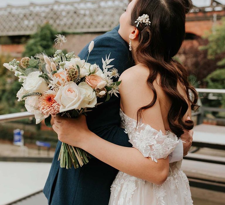 Bride in tulle lace off the shoulder dress and pearl hair accessory hugs the groom in blue suit