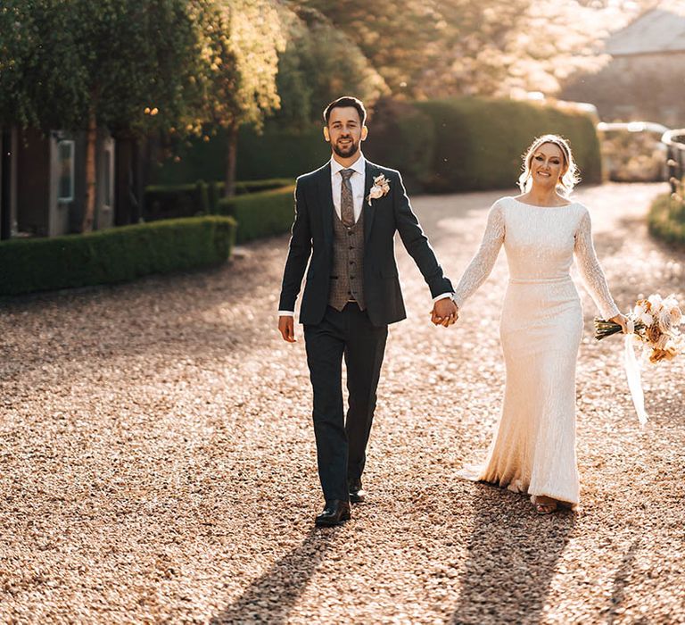 Groom in blue trousers and suit jacket with paisley tie and grey waistcoat holding hands with bride in high neck and long sleeve beaded wedding dress