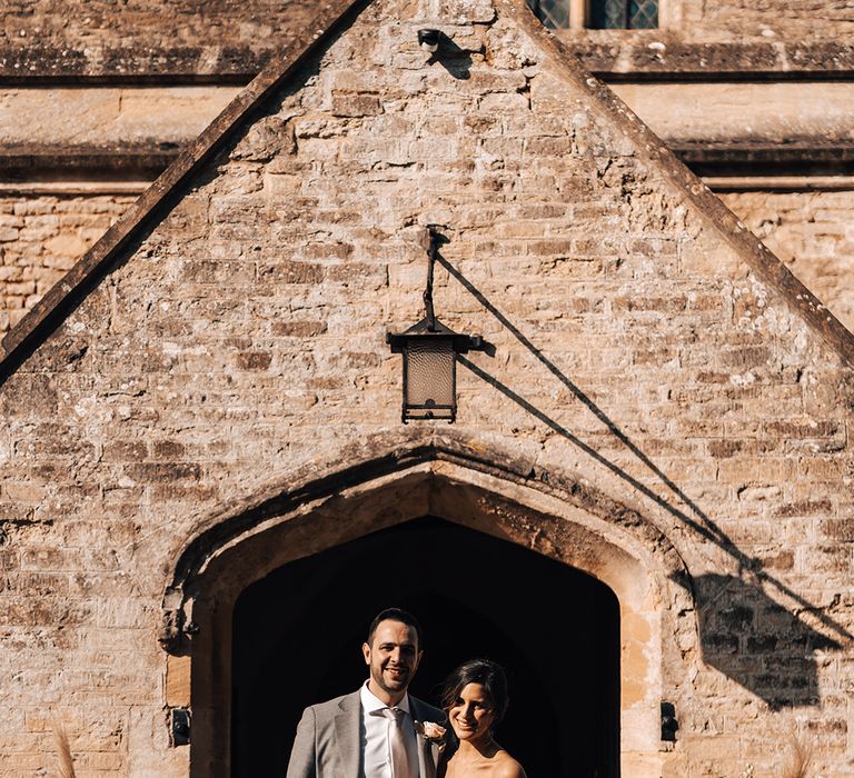 Bride in floral lace off the shoulder wedding dress with groom in grey suit standing outside wedding venue